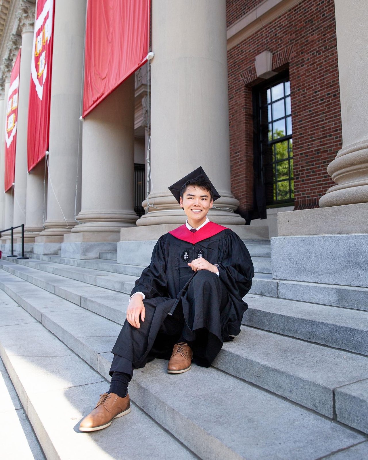 John Zheng sitting on the steps of Widener Library at Harvard in graduation regalia